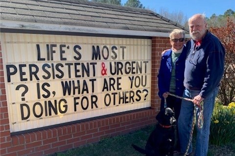 Man and woman with dog stand beside a sign about volunteering